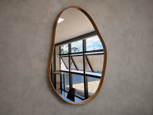 Espelho orgânico Nara com contornos assimétricos e moldura em madeira natural, pendurado em parede, refletindo uma janela com persiana